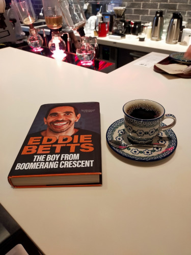 The photo is of a white counter with the hardcover book featuring a photo of Aboriginal footballer Eddie Betts smiling. His black hair is short & he has a fuller moustache & a sparse beard  To the right is an ornate blue & grey flowerly coffee cup & saucer of black coffee. A silver spoon is perpendicular in front. In the distance, 3 siphon coffee apparatuses can be seen, the middle bulb of water being bunsen burnered, its upper urn is full of coffee grounds