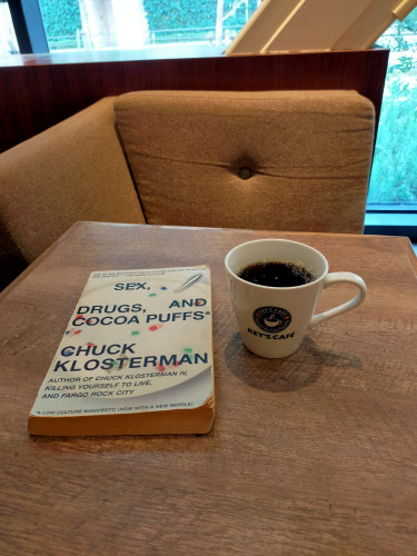 Photo is of the inside of the cafe  The white paperback book has an image of a bowl of milk with pill capsules in it instead of cereal. To the right is a white mug of black coffee with KEY'S COFFEE and a blue circle icon with a white mug of coffee in the center. Brown cushions of the chair opposite can be seen. Daylight can be seen out the window.