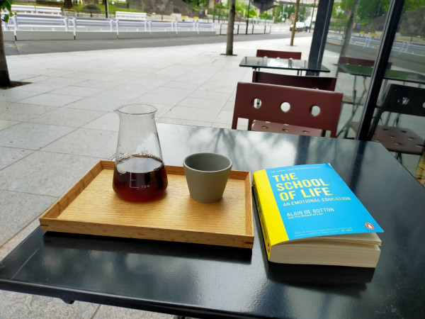 Photo is outside of a cafe. There is a square black metal table. The turquoise paperback book with a yellow spine is on the table. To the left is a wooden rectangle tray with a small greyish green cup. Further to the left is a beaker of black coffee. A vast sidewalk and a street can be seen in the distance