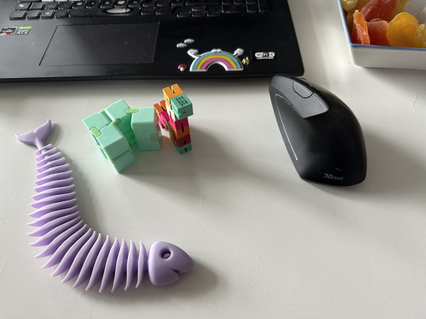 A photo of a desk with colorful fidget toys on it. You can also see a corner of an open laptop with pan rainbow and a few smaller stickers on it, a square bowl with some candied fruit and a vertical mouse
