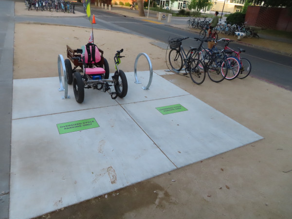 Two circular loop racks bolted to a large concrete pad. A green sign on the ground in front of each reads "Oversized Cycle Parking Only". I have parked the quadricycle in between the two racks so people can see that it's not wide enough for two quadricycles to park nor two tricycles, nor two handcycles. It is still an installation where the quad could only fit on the ends unless no one had arrived yet and I wanted to take up two places instead of one.

A rack of the inaccessible "lightning bolt" type is nearby.