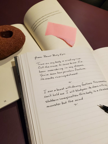 Handwritten excerpt from poetry collection Beast Body Epic by Amanda Earl - book is in the background, held open by a piece of red brick, with a torn pink post-it note marking the page, and an uncapped black pen rests on the notebook page