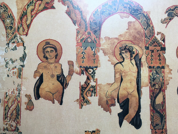 The remains of a colourful tapestry depicting Ariadne and Dionysos. Both are fully nude and both are depicted with a halo around their head. Dionysos holds a kantharos cup in his right hand and touches the top of his head with the left. That gesture and his facial ecxpression make him look a bit confused.