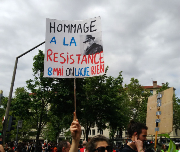 Une personne, dont on voit le haut du visage, tient dans son bras droit une pancarte : "Hommage à la Résistance - 8 mai on lâche rien".
A l'arrière des manifestants, un feu de circulation, des arbres et des bâtiments.
Le ciel est gris et nuageux.