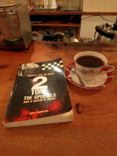 The photo is of a coffee shop counter on which is a black paperback book. To its right is a ornate cup of black coffee on a white saucer with red ringed rim. Various used french presses, cups, & other coffee shop implement s can be seen in the distance