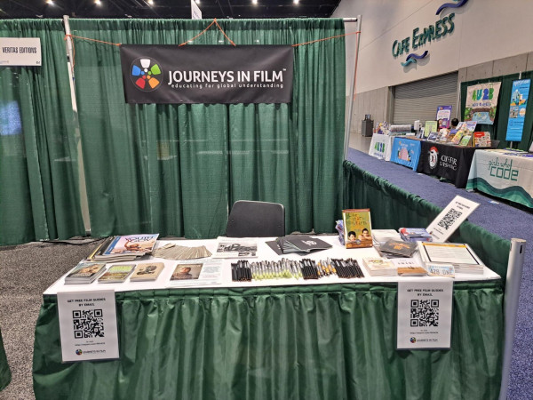 Photo from The Library Marketplace exhibition hall at the American Library Association Annual Conference. The Journeys in Film booth has a table covered in cool stuff, plus signs with QR codes to sign up for the newsletter and request film guides by email. 