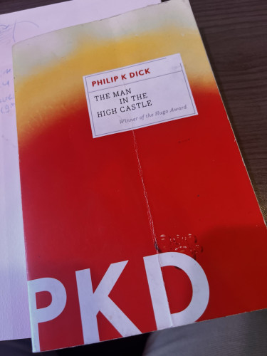 A book cover of "The Man in the High Castle" by Philip K. Dick. The cover features a gradient of yellow to red with large white letters "PKD" at the bottom. The book is positioned on a table with part of a