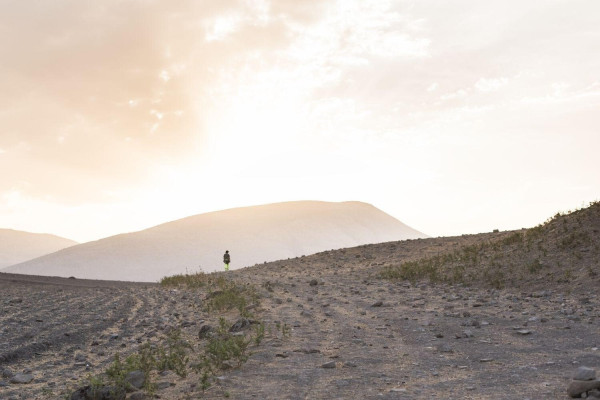 A single person walks in a barren, mountainous landscape. The sun is low and bathes the scene in warm light. 