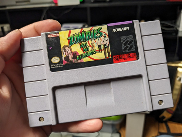 A photo of the SNES cartridge for "Zombies Ate My Neighbors". On the label, the game's title is overlayed over a frightened woman and a trio of zombies stumbling toward her