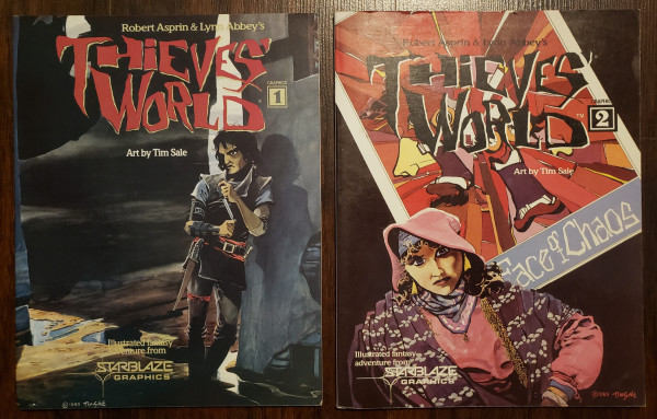 Thieve's World Graphics volumes 1 and 2. 
Volume 1 depicts a dark-haired man leaning on an old wall before a dark alley. He looks sinister and contemplative. 

Volume 2 has an inset image in the background that appears to be a tarot card, labeled "I" with an image of many mouths against an orange chaotic field, with the caption "Face of Chaos." 

In front of the card is a person in pink hooded robes with a purple shawl draped over their shoulders. They might be a mystic or card reader.
