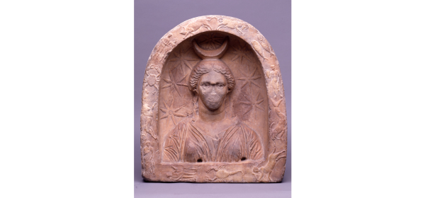 Reddish marble carved votive relief of Selene: a female bust to the front in an arched niche, draped in a sleeved chiton, and with her hair parted in the middle and brought down over her ears, nose now missing, surrounded by a crescent on her head, seven stars in the field around and the signs of the zodiac in low relief; inscribed beneath with an unitelligible Gnostic formula and noted in the catalogue that from its position the inscription appears to have been an afterthought.