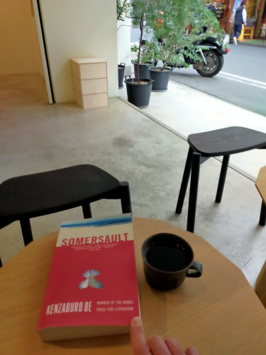 The photo is of seating outside a cafe. The mostly pinkish red cover with an upside down down of the paperback book is next to a black mug of black coffee on a brown coffee table.  A motorcycle & red bicycle can be seen in the alley in the distance