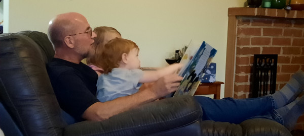 Grandpap sitting in a recliner with 2 children on his lap. One is reaching to point to the book he is reading aloud to them. Both are giggling.
