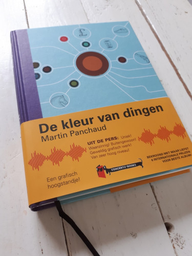The Dutch edition of the book "The Color of Things" is displayed on a white wooden table. It is a beautifully bound hardcover book with a reading ribbon. On the blue title page, there is a brown circle in the centre connected to other circles with white thin lines.