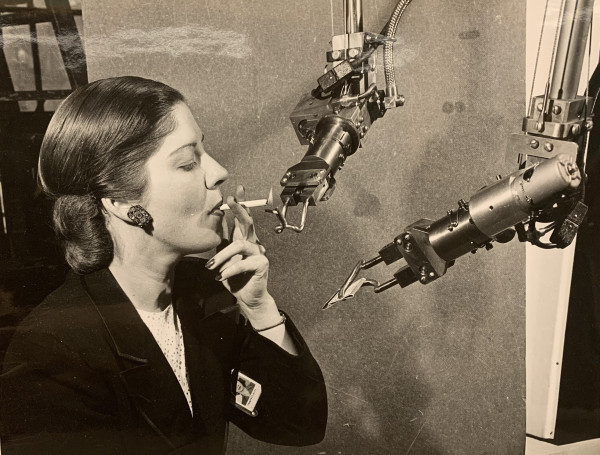Square black and white image with woman employee in dark sports jacket on left and two mechanical robotic arms on the right. General Electric was showing off various forms of robots in the 1950s. Here, we see mechanical hands, striking match & lighting a cigarette of this GE employee.