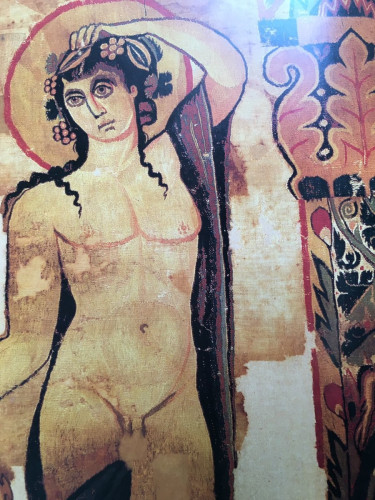 Photo of a tapestry depicting the god Dionysos with grapevine in his hair, his dark hair flowing down over his shoulders, and a purple robe hanging from his left arm that he has raised to touch his head in a pose typical of his brother Apollon. The god is naked, with little dots painting a circle around his nipples.