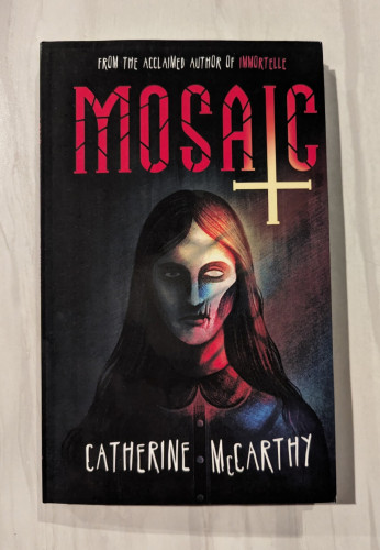 Book cover of MOSAIC by Catherine McCarthy. An illustration of a person is depicted with the shadow of colored stained glass shining on them. Half of their face is a skull. The title is above the person with the "I" in "mosaic" turning into an upside down cross.