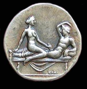 Silver spintria depicting a woman and a man atop a bed or couch. He is reclinig while she is giving him a handjob.