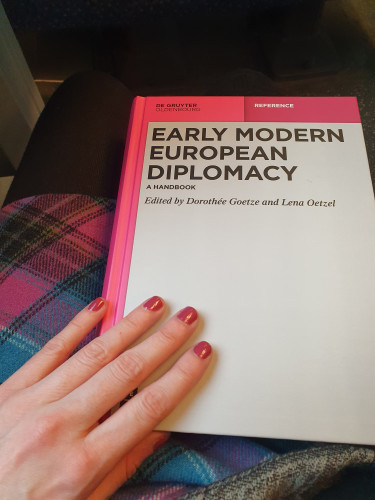 Pink/grey book (Early modern European Diplomacy. A Handbook, ed by Dorothée Goetze and Lena Oetzel) lying on a modern pink tartan skirt. You also see a hand with pinkish nail polish. 