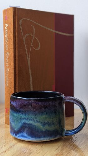 A picture of a very pretty mug positioned in front of a book of classic American Short Stories. The mug is horizontally banded with dark blues, greens, and purples in a way that evokes the Northern Lights.