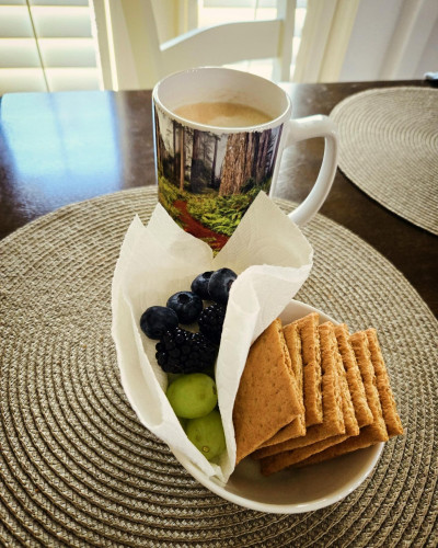 Coffee mug with forest photography, bowl of fruit and Graham crackers on beige woven placemat.