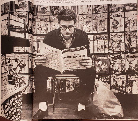 A picture of a large black and white photo across two pages of a February 2, 1962 "LIFE" magazine. In the photo, a young man with short dark wavy hair, wearing thick rimmed glasses, a light-colored collared shirt, a dark cardigan sweater, dark trousers with cuffs, white socks, and black Oxford shoes, sits crouched forward on a padded seat with chrome legs, reading a thick magazine, as follows:

"128 pages of Tax Information that can save you money. NEW EXPANDED EDITION. $1 America's Favorite INCOME TAX GUIDE. The Tax Digest, Inc. New York."

Most of the, then New, comics have a cover price of twelve cents. Titles and characters visible in the photo include:

Woody Woodpecker, "GIRLS' ROMANCES," BATMAN, "Walt Disney's DONALD DUCK ALBUM," Dennis the Menace, "SUPERBOY," "Playful LITTLE AUDREY," "LIFE with Archie," King Leonardo, Barbie, Looney Tunes, Baby Huey, Archie Comics "LAUGH," "MYSTERY IN SPACE," "WORLD'S FINEST," "Mickey Mouse," "Huckleberry Hound," Bugs Bunny, "UNCLE $CROOGE," "Adventure Comics," "Archie's MAD HOUSE," "SUPERMAN'S PAL Jimmy Olsen," and more.