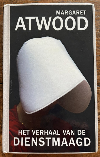 Omslag van een boek getiteld "HET VERHAAL VAN DE DIENSTMAAGD" door Margaret Atwood, met een persoon in het rood met een witte muts tegen een zwarte achtergrond.