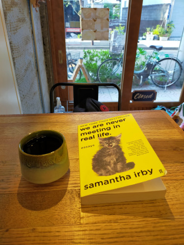 The photo is inside the cafe where you can see out the front window an electric bike against greenery along the sidewalk. In the foreground is a wooden table with the yellow paperback book with a wet grey growling cat illustration. To the right is a round tea-like ceramic cup of black coffee.