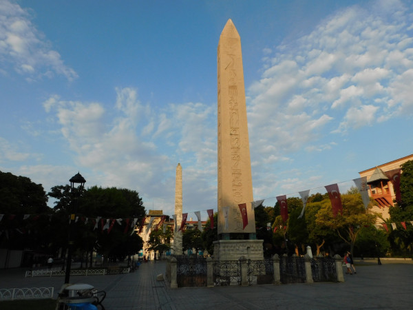 The obelisk from a distance, rising about 33 meters above a meter-tall square stone pedestal. The pedestal is at ground level, but there is a pit dug around it and more supports below it. There is a railing around the pit.