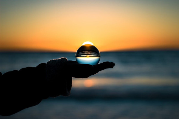 Photograph of an outstretched arm, holding a crystal or glass ball in an open palm. It is shot against a sunset over the sea, the ball placed right where the sun would be.