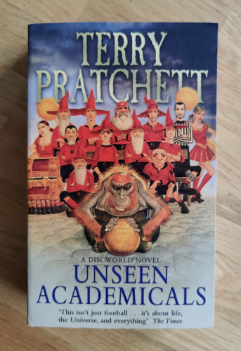 A soft cover copy of Unseen Academicals by Terry Pratchett.  A motley crew of wizards, most in pointy hats, wear red football kits.  There is also an orangutan holding a football, a referee, and a cheerleader.