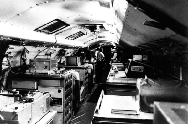 Black and white image of the inside of a plane's fuselage that is set up with computer equipment in the foreground in rows against the edges of the plane with a row to walk in and access equipment down the middle. A few men stand in the background.