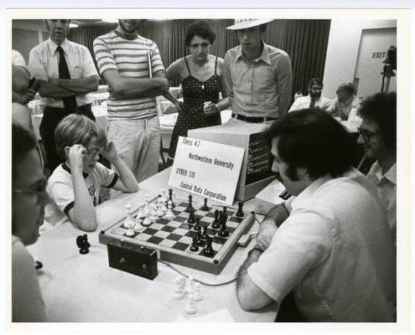 Black and white rectangular horiz. orientated photo. A child plays chess against a Control Data Corp computer program at a demonstration at the Palmer House in downtown Chicago, IL, 1979. There is the chess board, a white tablecloth and a group of adult onlookers the boy is touching both sides of his glasses and is in deep concentration. There is also a timer with two buttons in the foreground.