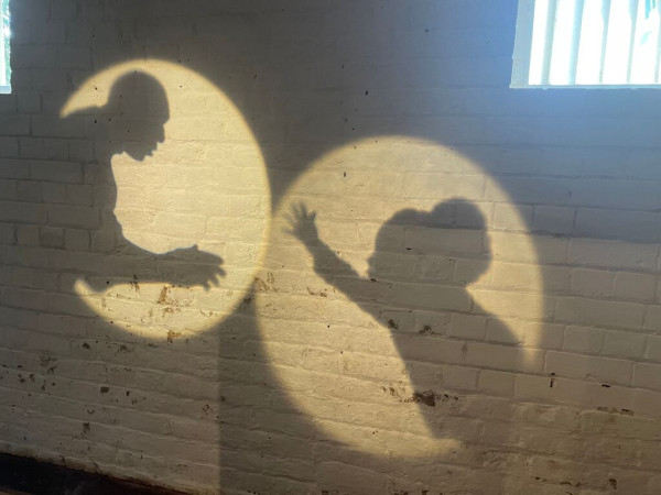 Shadows of a man and child reach out for each other. Both figures are projected onto a white brick wall, light comes through a barred window in the top right corner.  