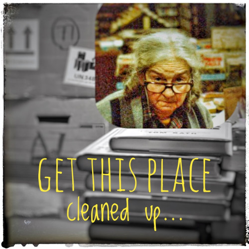 A black and white photo of boxes piled high with a stack of books in the foreground and slightly to the right of frame. Above the books is a color inset photo of an elderly woman peering sternly over her glasses into the camera. A caption below her reads, "Get this place cleaned up..."