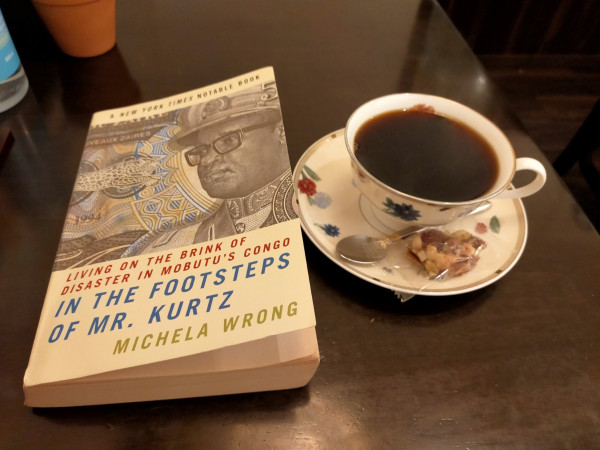 The photo is of a table on which is the paperback book mentioned in the toot. It is white with an image of an old banknote with the late Black dictator of Zaire & the Congo, Mobutu Sese Seko in a general 's hat & Buddy Holly black glasses. To the right is a white cup & saucer of black coffee, with a few colorful flowers designed on the cup & saucer. On the saucer in front is a silver spoon & a plastic wrapped nut square bar.