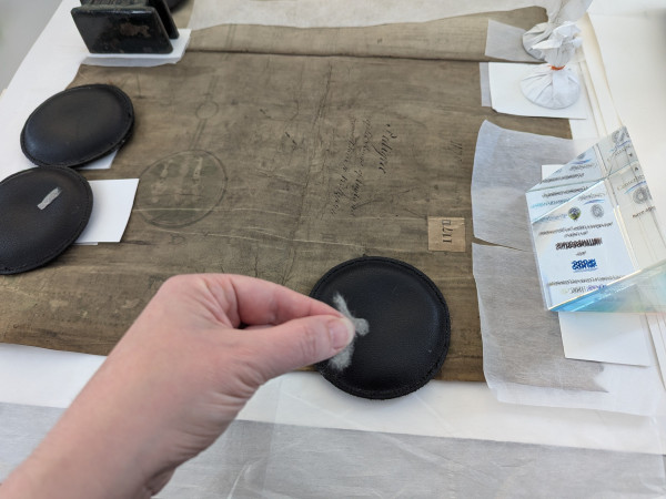 Dark brown from engrained dirt parchment roll. Multiple areas repaired with item held down with dark coloured circular weights. In the foreground, a shaped piece of tissue held with a left hand.