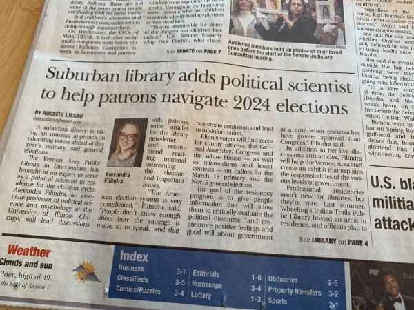 A photo of the front page of a newspaper. The highlighted article has the headline 'Suburban library adds political scientist to help patrons navigate 2024 elections'. Accompanying the article is a small photo of a woman in glasses, captioned Alexandra FIlindra.