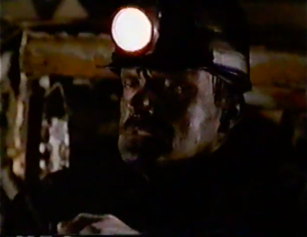 A man in a miner's helmet. He's sitting in a dark, small space, and his face is covered in dirt. His expression is grim