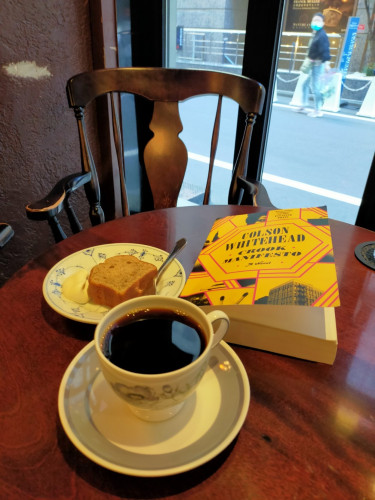 The photo is of a dark brown round wooden table inside a coffeehouse. In front is a mug & saucer of black coffee. The yellow paperback book is behind it, as is a white plate with a slice of pound cake with cream. And old style wooden chair is behind the table & an alley view can be seen our the window where a blurry masked Asian woman can be seen looking at the canera