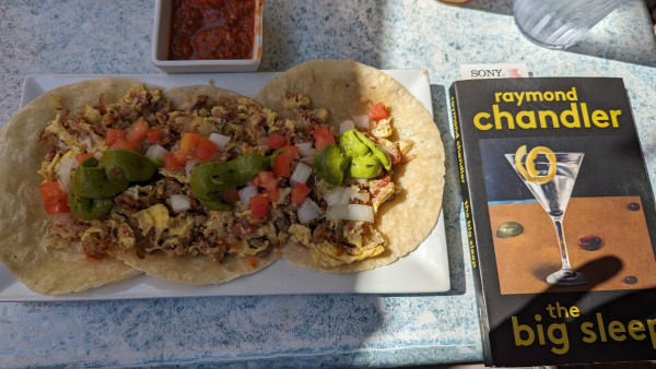 Three breakfast tacos on a platter with some salsa. To the right is a paperback copy of The Big Sleep by Raymond Chandler. The cover shows a picture of a martini with a twist of lemon peel.