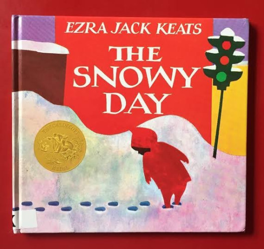 Cover of children's picture book, "The Snowy Day" by Ezra Jack Keats. There is a Caldecott Gold foil emblem on the left. The title and author are in white on a bold brick colored square in the center flanked by snow lining the top of a darker brown wall to the left, and a goldenrod wall to the right. A traffic signal tips just slightly. The foreground is a young black child in a red snowsuit turning and looking behind himself at his footprints in the snow. More snow heaps up behind him. 