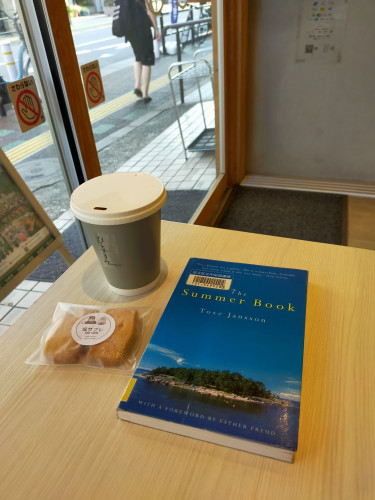 Photo is inside the cafe against the window sliding doors. Outside the window a woman in a black dress can be seen walking on the sidewalk. On a light brown table in the cafe is a blue paperback library book with a photo of an island off the north of Finland. To the left is a paper coffee cup with a white paper lid and a gray body with says ひとやすみ vertically. In front of the cup is a plastic covered pair of rectangular cookies.