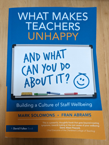 Front cover of "What Makes Teachers Unhappy and What Can You Do About It?" by Mark Solomons and Fran Abrams. It is blue, orange and white with the second half of the title written on a whiteboard with a smiley face drawn next to it. 