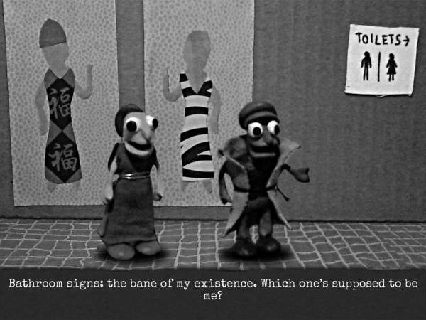 Screenshot of claymation scene. The Background is a simple cardboard wall with two posters of figures in long dresses. On the wall to the right is a sign saying "Toilets ->" with two traditionally gendered symbols for "man" and "woman" below.
In front of the wall stands two human clay models. The one on the left is a girl wearing a simple long dress. Her facial features is exaggerated with big eyes, nose and mouth.
The one on the right is a gender-ambiguous person wearing a clay cap, a piece of fabric wrapped around them like a coat and tied at the waist, and pants. They also have exaggerated features.
Their arm is out indicating they're talking at the moment.

The text below says "Bathroom signs: the bane of my existence. Which one is supposed to be me?"