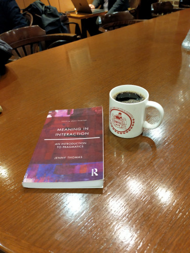 The photo is of the purple paperback book on a brown table. A white mug of black coffee with the red Saito circular medieval logo on is to the left. A man can be seen using his laptop sitting in the distance but you cannot see his face