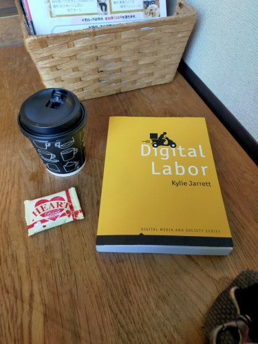 Photo of a wooden bench on which is the yellow book with a black illustration of a scooter delivery driver heading right. To the left is a small black coffee cup & lid, the cup having white outlined images of cups. In front of it is a white plastic packaged chocolate that says HEART