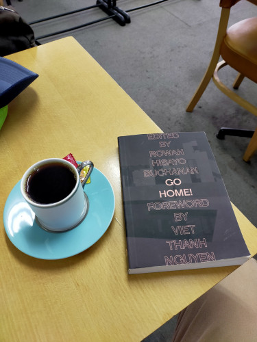 Photo is above a yellow square wooden table. The black paperback book is on the table. To the left is a turquoise saucer on which is a white mug of black coffee