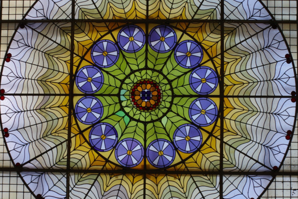 Photograph of a stained glass window. Window is circles within circles; outer largest circle has spiderweb-like pattern between spokes, inner circle has blue small circles in a wheel pattern. Colors are green, orange, blue and white.