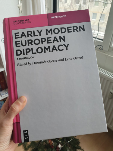 Copy of Early Modern European Diplomacy. A Handbook, by Dorothée Goetze and Lena Oetzel (de Gruyter). You see the pink/grey cover and a hand holding it.